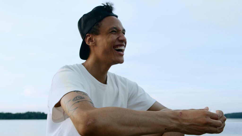 A young man in a cap and t-shirt laughing joyfully by a serene lake.